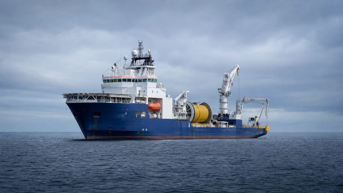 Large white ship with a bulky stern winch on open gray sea, laying cable from a spool.